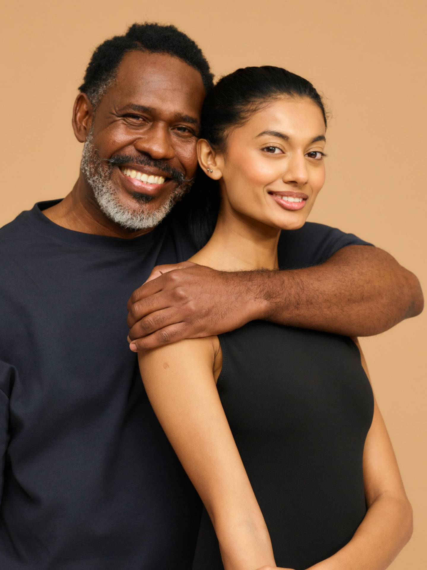 An older man and a younger woman smiling warmly together against a tan background